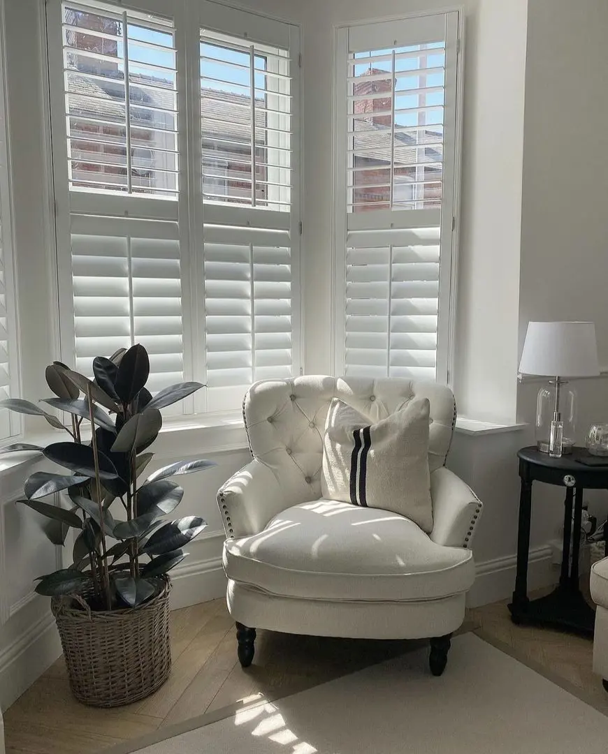 Splayed living room bay window wooden shutters with cream armchair